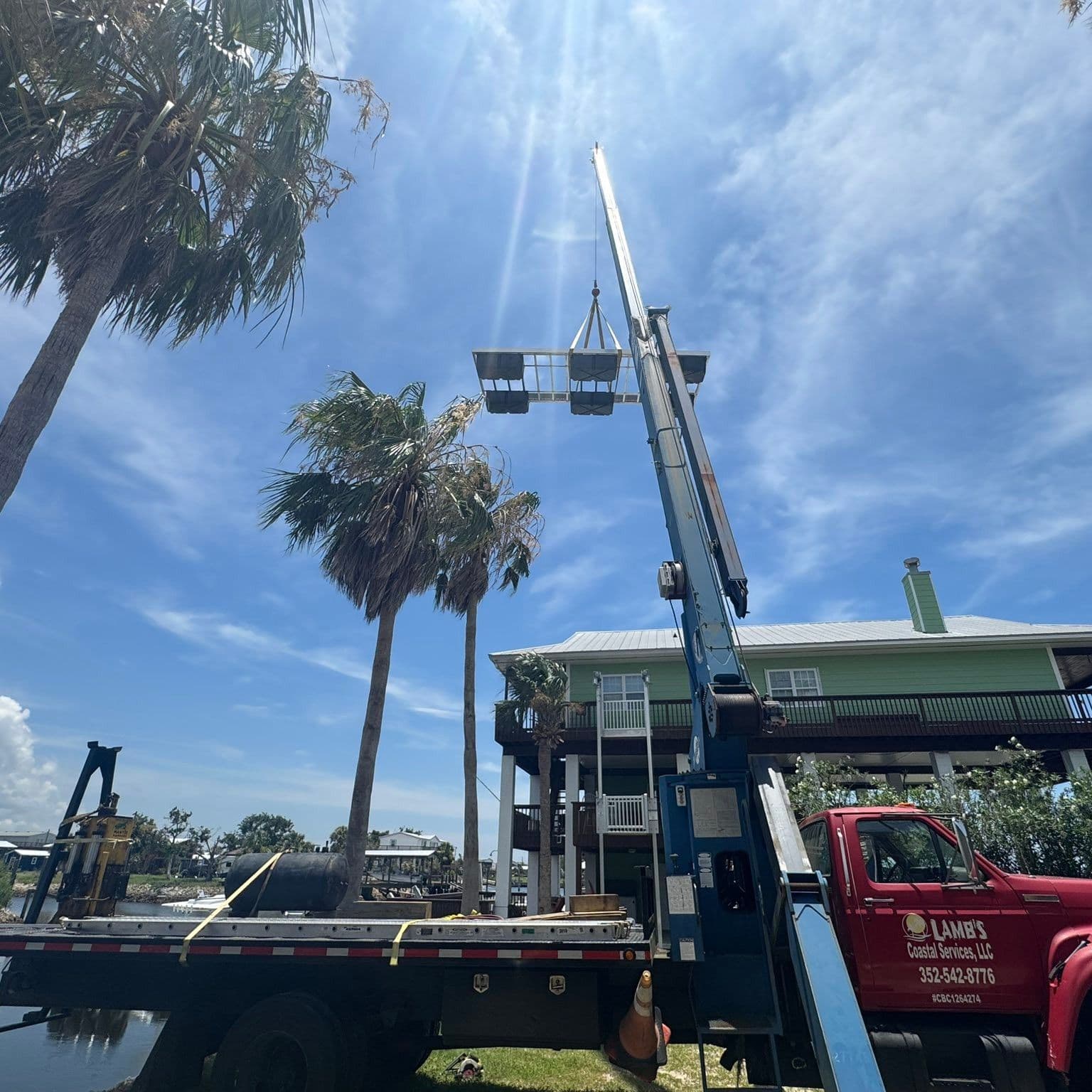 Red Lamb's Coastal Services crane truck, with the company name, phone and license number on the door, hoisting materials beside a raised home
