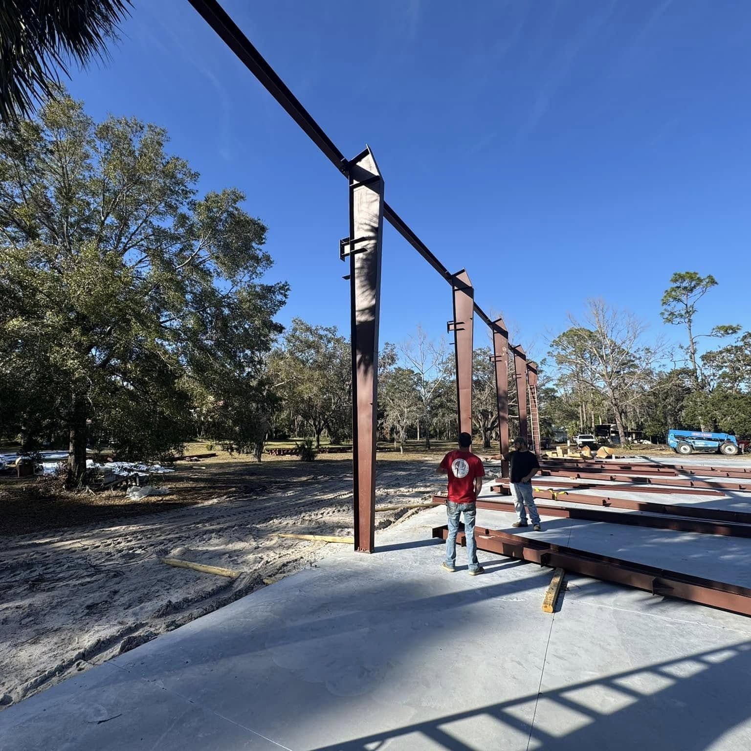 Red-primed steel columns and beams erected on a concrete slab, two people standing nearby