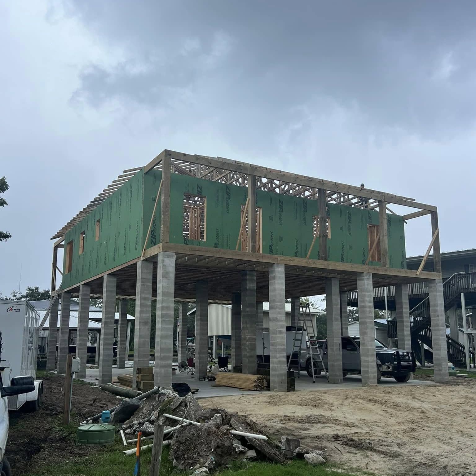 Raised home framed and wrapped in green sheathing on block piers under a cloudy sky, lumber and debris on the lot