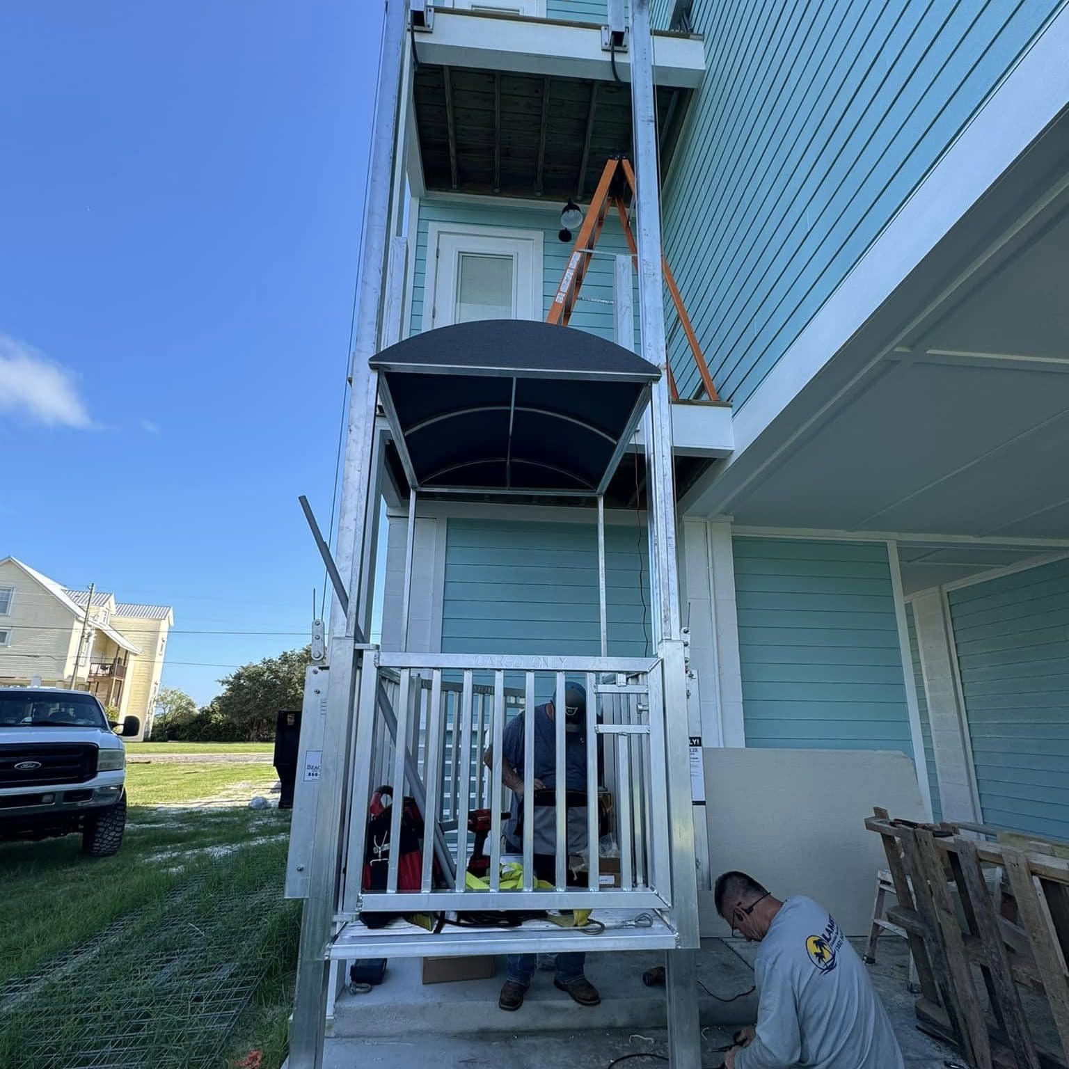 Cargo lift car with a black canopy at the base of a light blue raised home while crew members work on the install