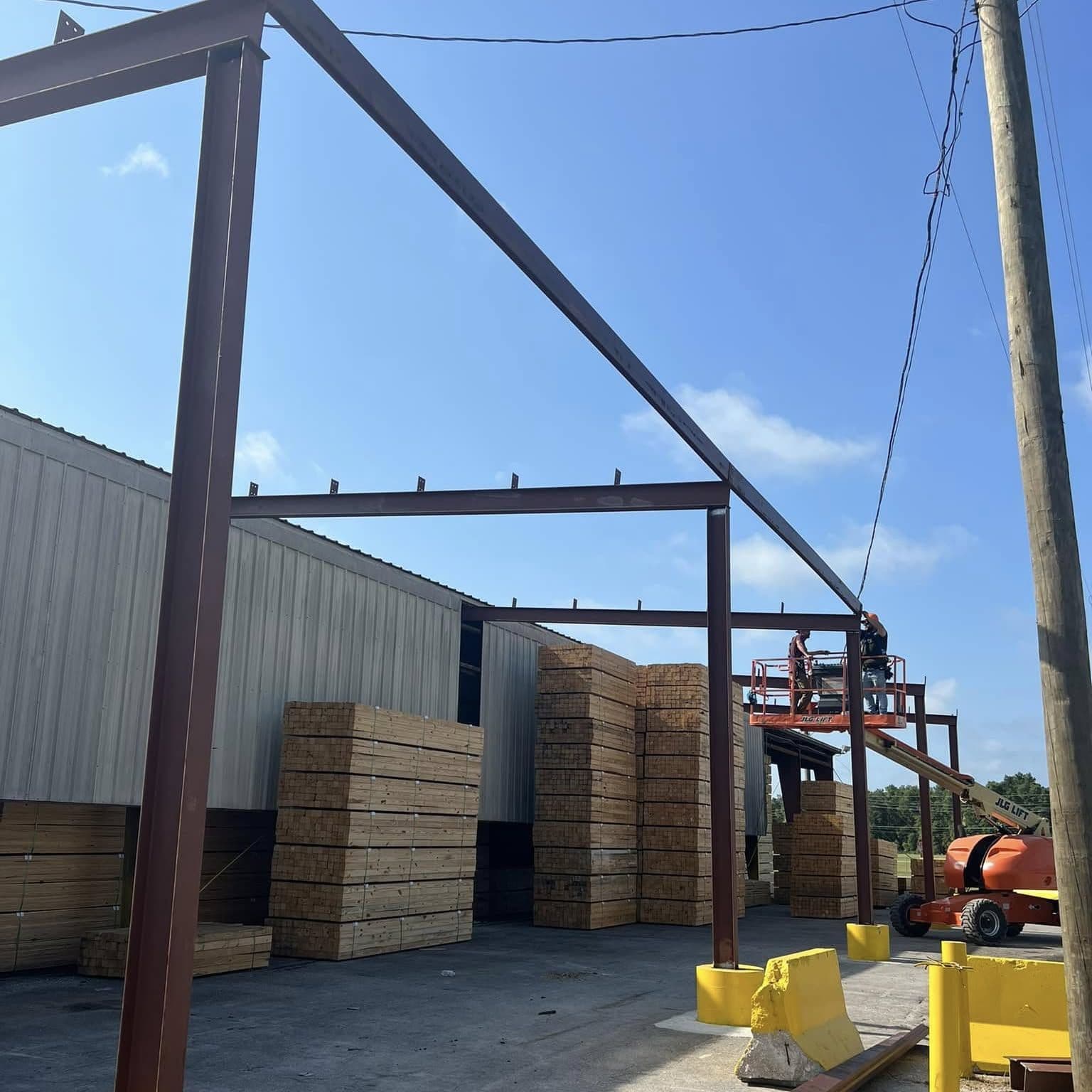 Red-primed steel canopy frame going up beside a metal warehouse with stacked lumber and a boom lift