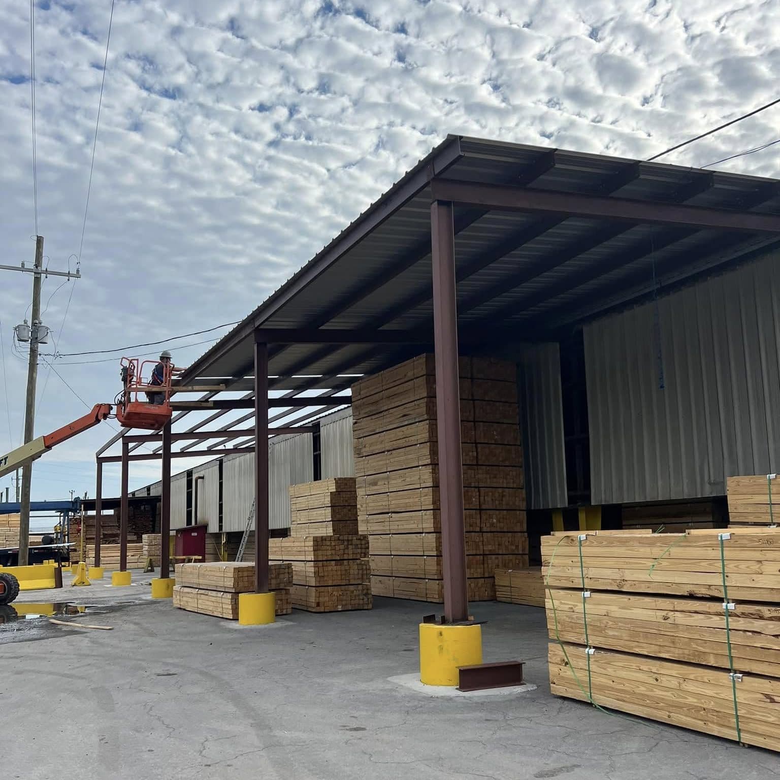 Finished steel canopy roof over lumber storage beside a metal warehouse, yellow bollards at the columns