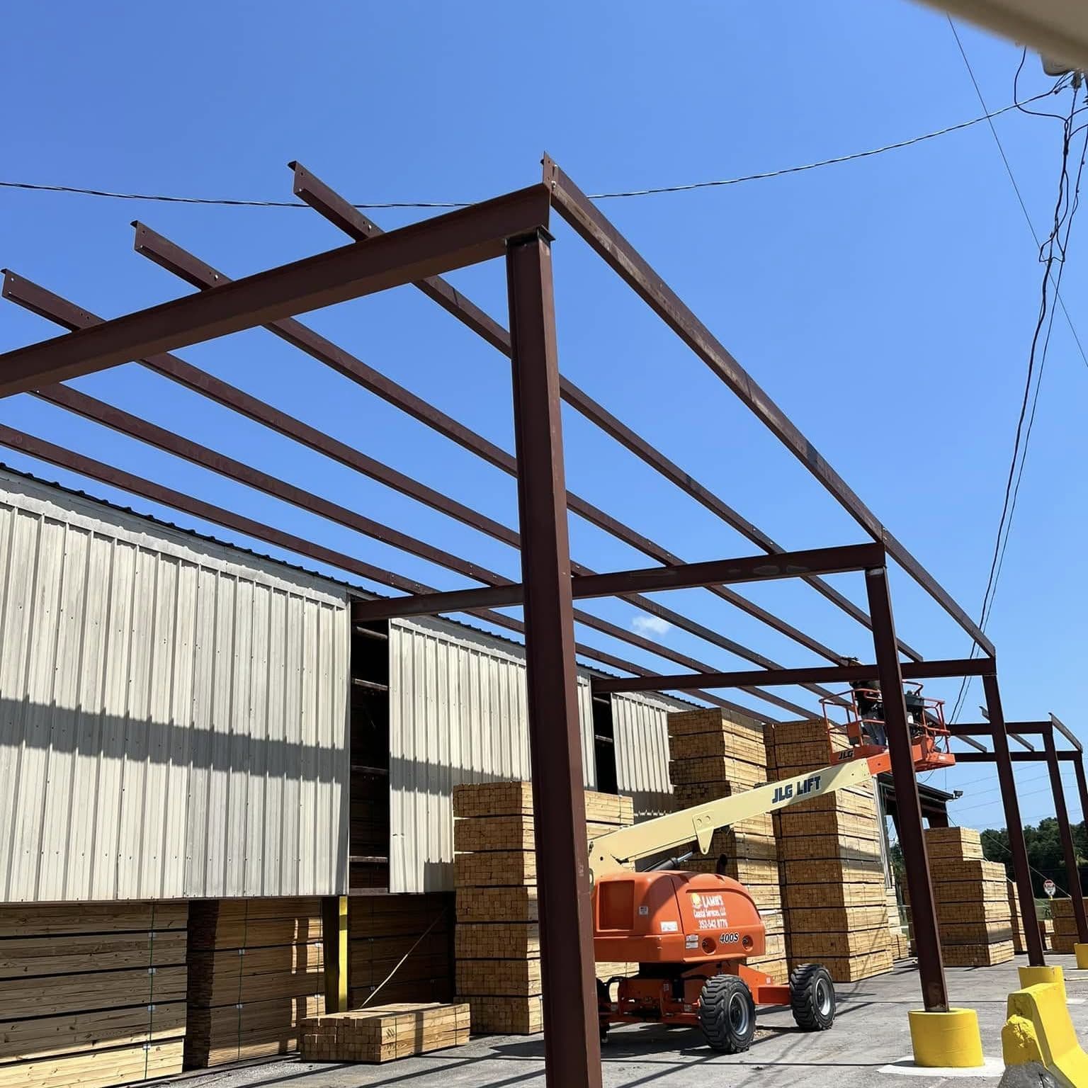 Steel canopy rafters and columns before roofing, with lumber stacks and an orange boom lift underneath