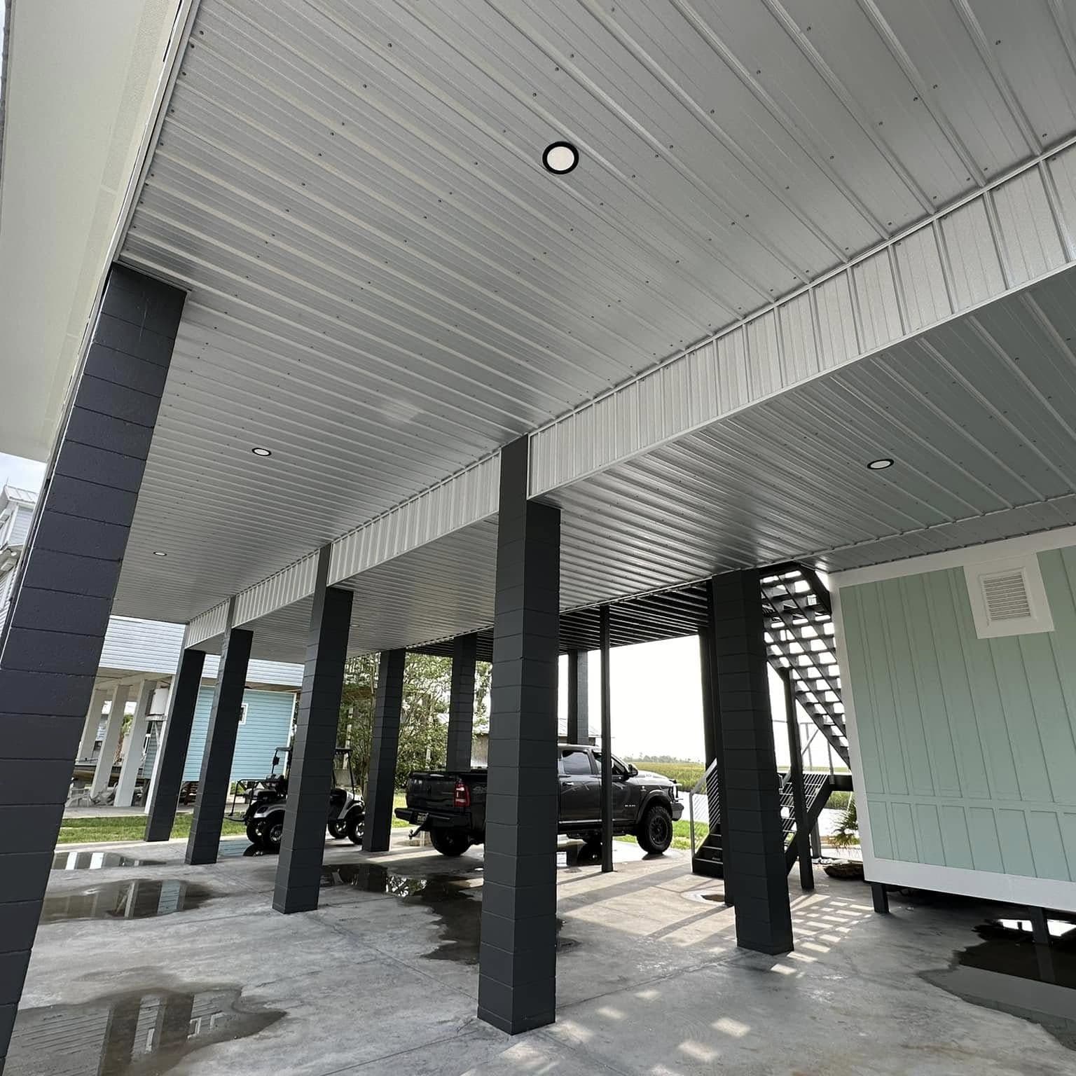 Covered parking under a finished raised home: white ceiling with recessed lights, dark columns and a concrete slab with a pickup truck