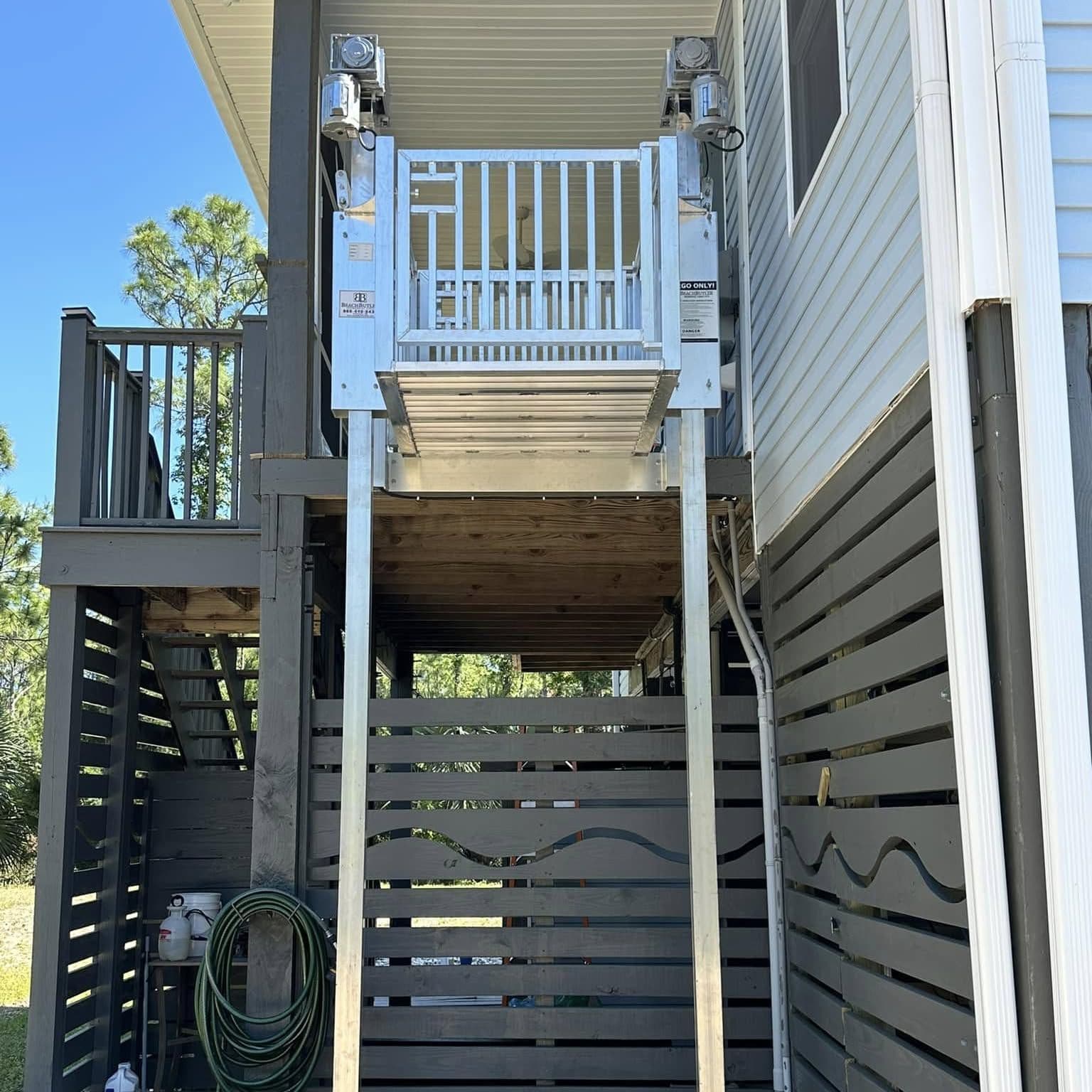 Cargo lift car at the upper landing beside a raised home's deck, with a dark slatted privacy screen below