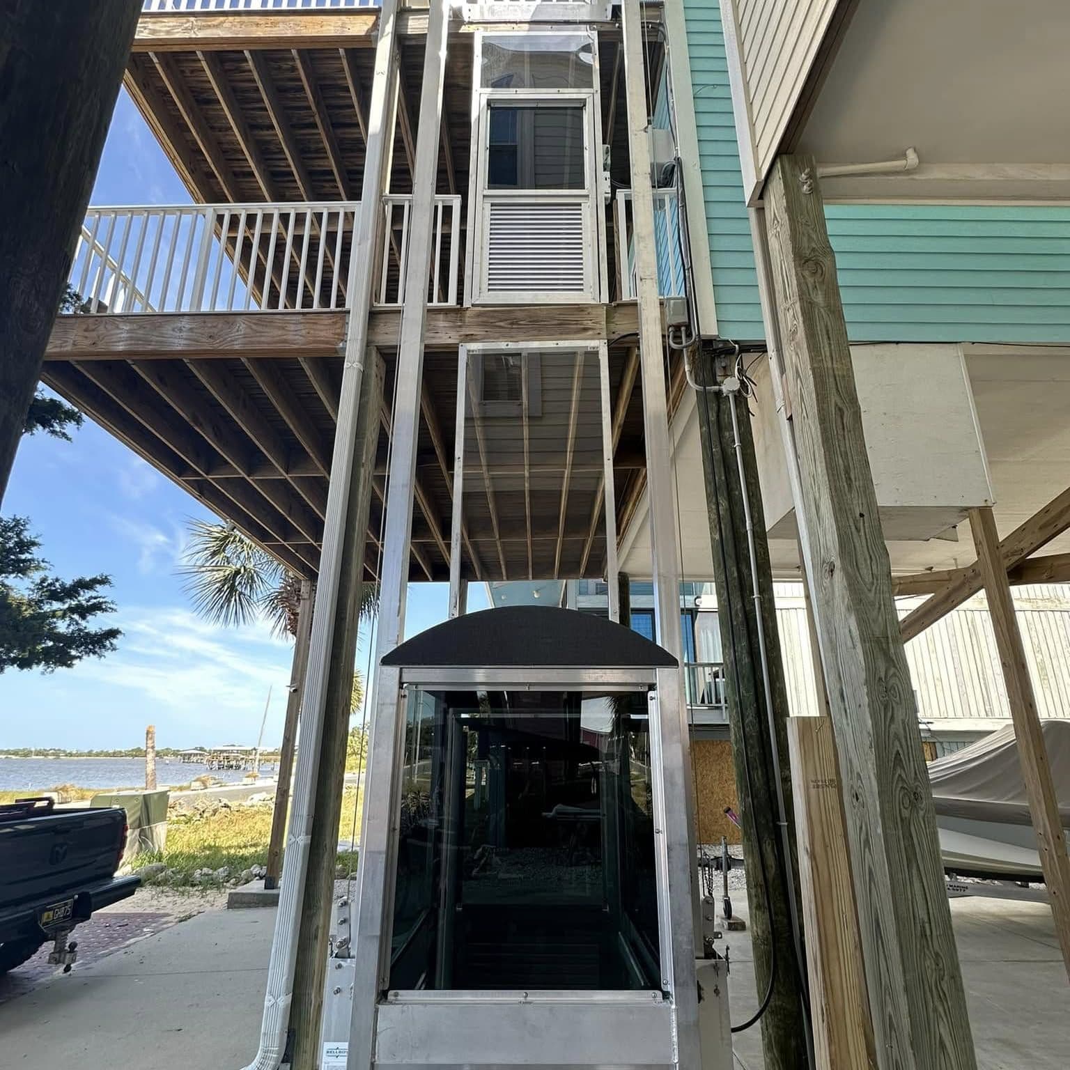 Glass-sided lift cab on aluminum rails under a raised waterfront home, rails running up to the deck