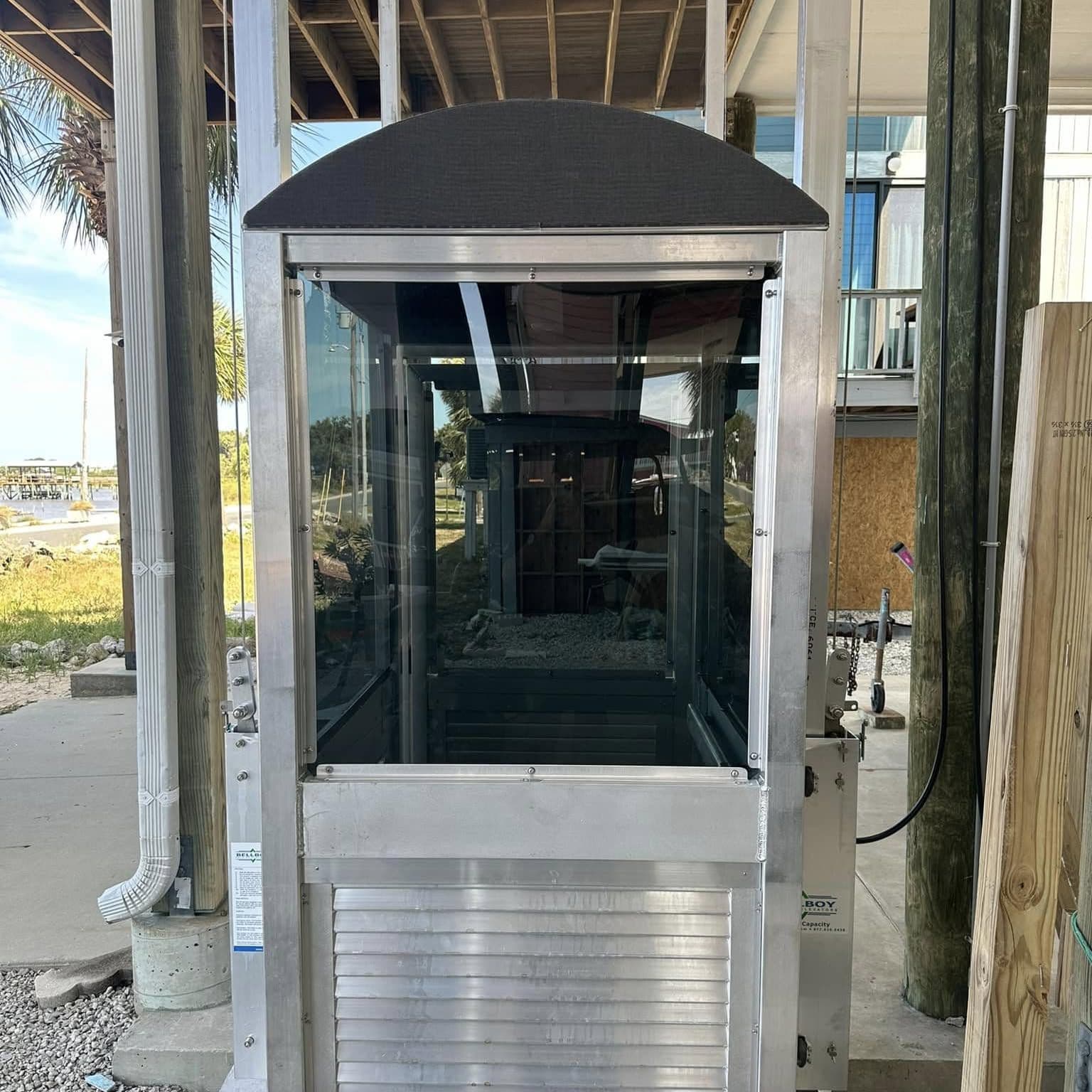 Enclosed glass-and-aluminum lift cab with an arched roof at the ground stop under a raised home