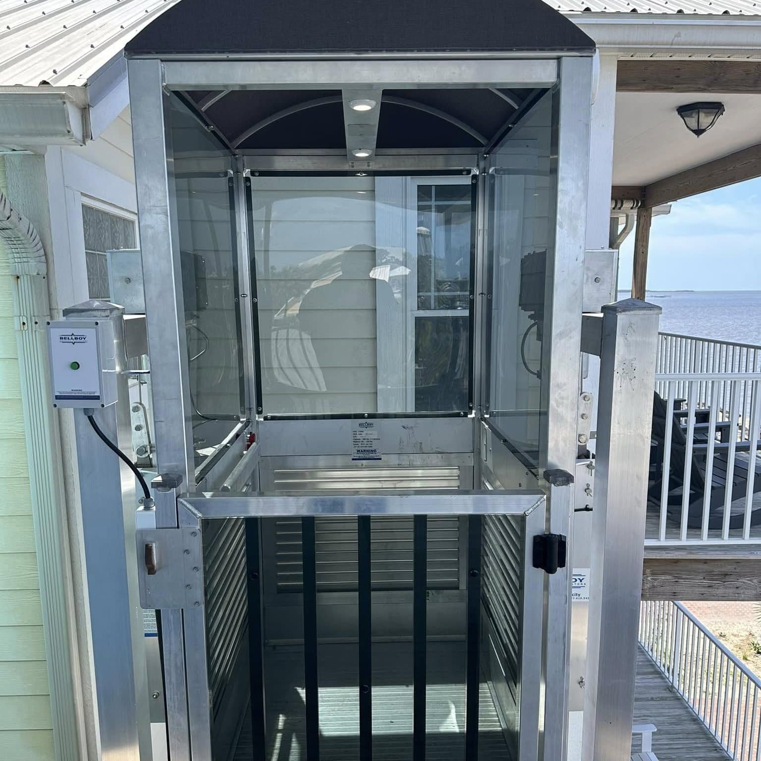 Front of a glass lift cab with a barred lower gate and a wall-mounted control box, waterfront deck behind