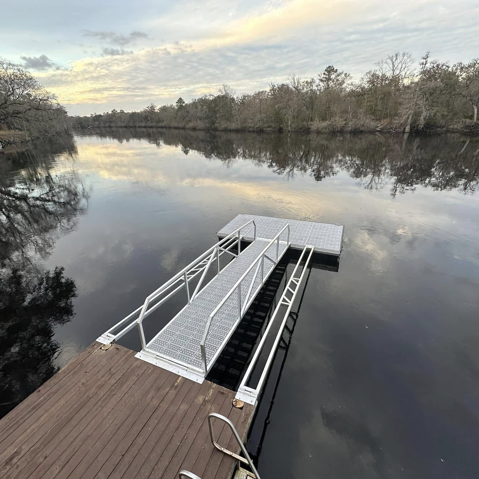 Aluminum gangway from a wood deck down to a floating dock on a tree-lined river at dusk