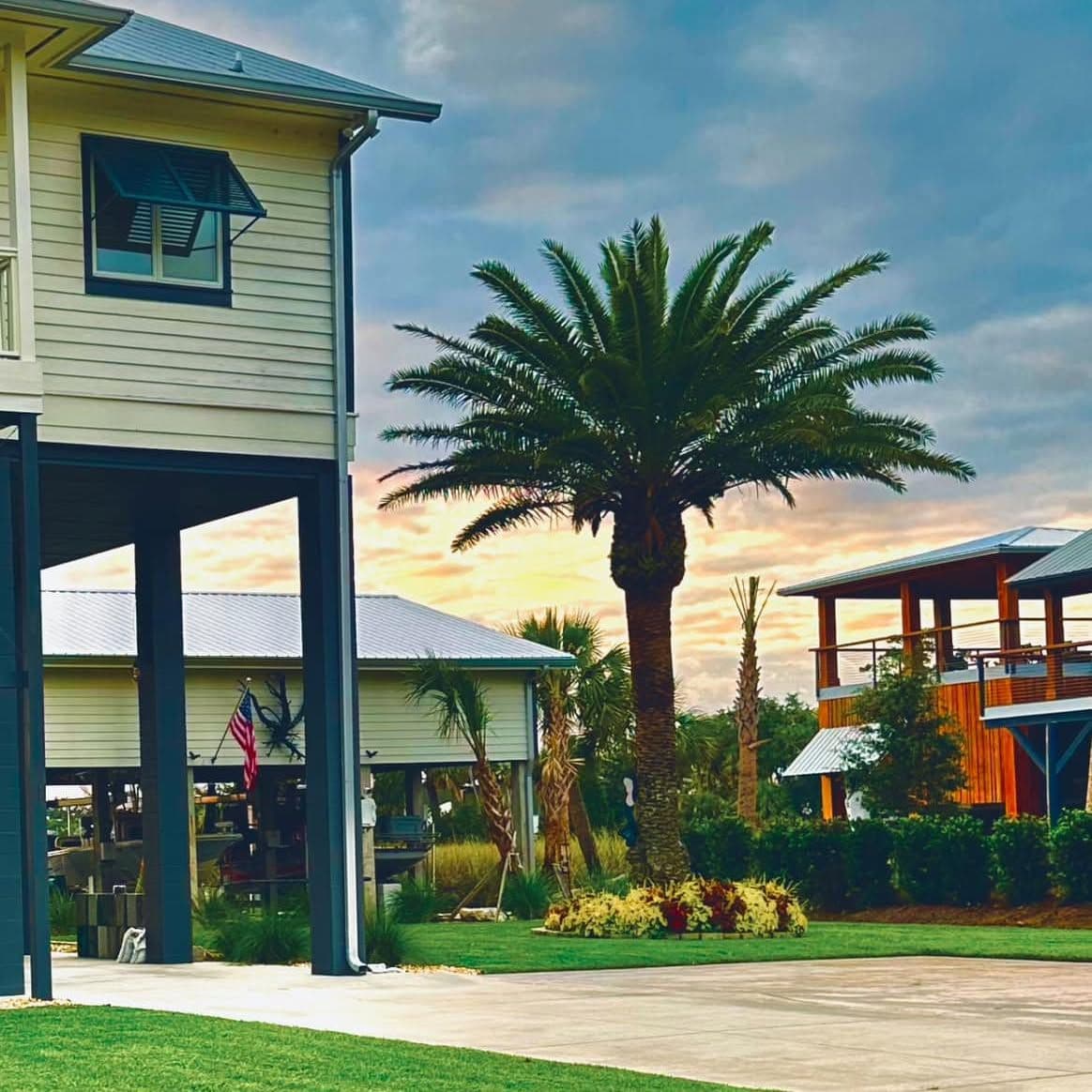 Raised home on dark blue piers beside a large palm tree, green lawn and shrubs, with a colorful sunset sky behind