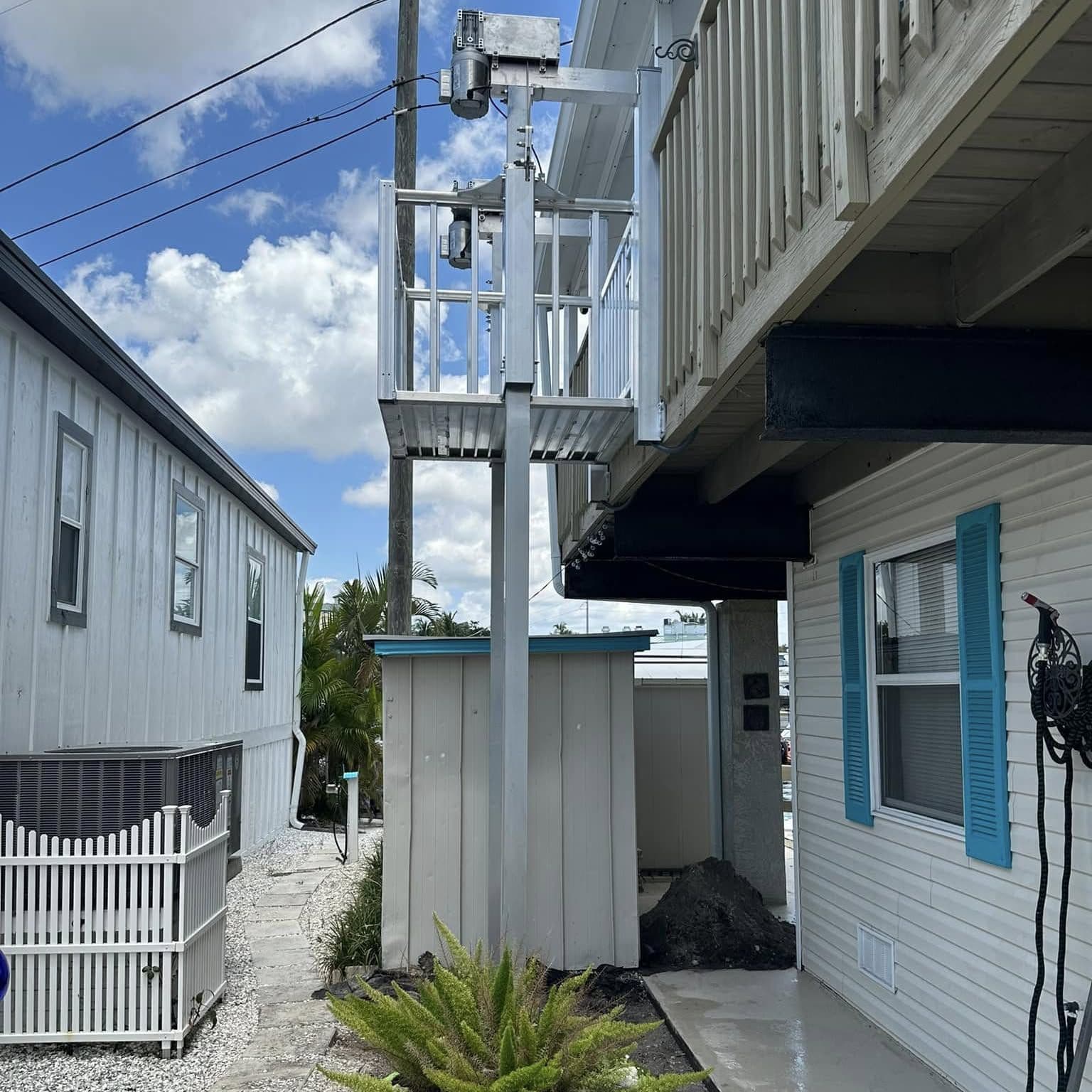 Cargo lift tower with a railed car at the upper deck along the side of a white home with blue shutters